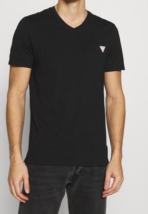 Mężczyzna w jednolitej czarnej koszulce z dekoltem w serek i małym trójkątnym logo na lewej piersi, w ciemnych dżinsach, stojący na jasnym tle.