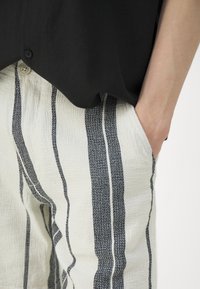 Person som bär vita byxor med vertikala svarta ränder och en svart skjorta, med handen avslappnat placerad i sidfickan på byxorna.