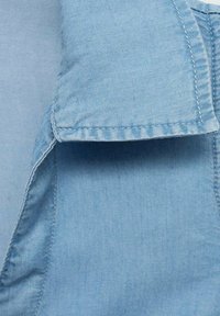 Tissu en denim bleu clair avec une texture lisse. Comprend un col et des coutures visibles le long des bords.