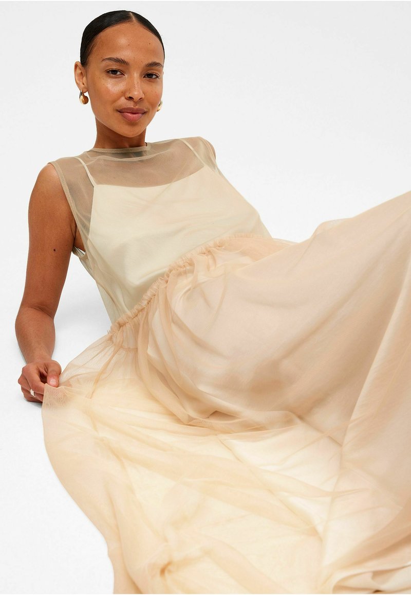 Lichtbeige mouwloos dress met een transparante overlay, een gathering in de taille en een vloeiende tule stof voor een zachte, luchtige uitstraling.