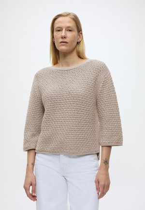 Vrouw met blond haar die een beige gebreide trui met textuur en witte broek draagt, staand tegen een effen witte achtergrond.