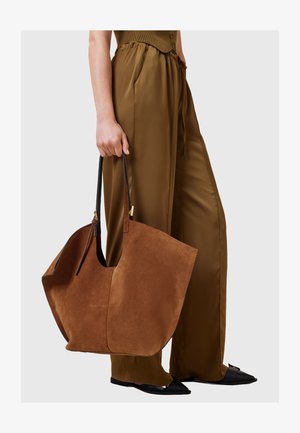 Pruun suede kott mustade nahkvarrukatega, millel on lai, lohkjas kuju ja minimalistlik disain, hoiab käes inimene pruunides pükstes.