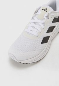 Chaussure de sport blanche avec une tige en mesh, comportant des accents noirs et trois bandes. Surface texturée et semelle amortie pour plus de confort.