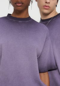 Deux personnes portant des hauts violets texturés à manches courtes assortis, l'une avec des boucles d'oreilles pendantes et l'autre avec un collier en argent.