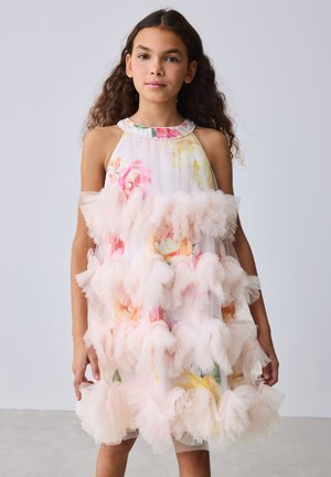 Jong meisje met lang krullend haar die een mouwloze witte jurk draagt met kleurrijke bloemmotieven en meerdere lagen roze ruches.
