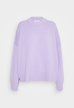 Henkilö, jolla on yllään laventelinvärinen ylisuurikokoinen neulepusero, musta nahkainen midi-hame ja mustat kiiltävät loafersit, kantaa suurta mustaa nahkaista clutch-laukkua.