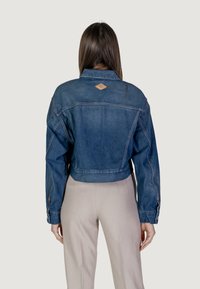 Giacca di jeans cropped di colore blu scuro con un'etichetta marrone sul retro, caratterizzata da un design rilassato e dettagli di cucitura visibili.
