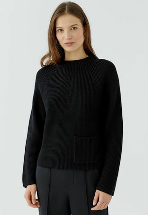 Vrouw met steil bruin haar, gekleed in een zwarte ribgebreide trui met een voorzak en zwarte broek tegen een effen lichte achtergrond.