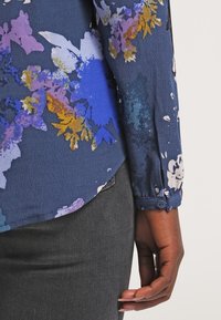 Marinblå blus med blommönster i livfulla lila och gula nyanser, lättviktsmaterial, långa ärmar med knäppta manschetter och en avslappnad passform.