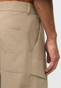 Κοντινό πλάνο του χεριού ενός ατόμου που ξεκουράζεται κοντά σε μπεζ cargo σορτς με μεγάλες τσέπες και κεντημένο κείμενο σε μία από τις τσέπες.