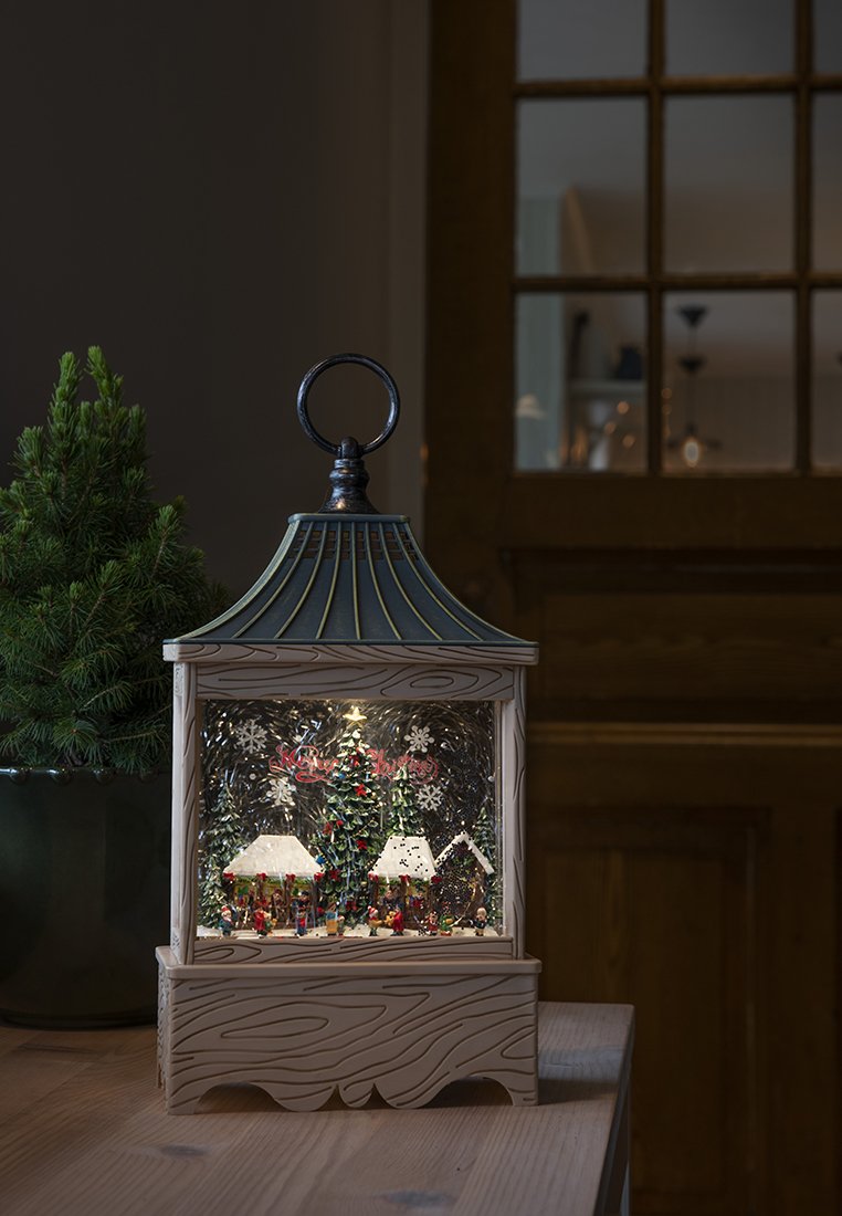 Farol decorativo con un mini pueblo navideño, árboles iluminados y figuritas sobre una mesa de madera junto a un árbol de hoja perenne en maceta.
