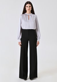 Seidene, hellgraue Bluse mit einem Ausschnitt in Form eines Schlüssellochs und gerafften Bündchen, kombiniert mit hoch taillierten, weiten, schwarzen Hosen. Schwarze spitze Schuhe.