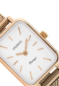 Montre rectangulaire dorée avec un cadran blanc, fins index en or et un bracelet en métal tressé. Équipée d'une couronne de remontage sur le côté.