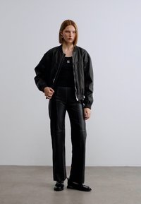 Μαύρο δερμάτινο μπουφάν τύπου bomber με φερμουάρ μπροστά, συνδυασμένο με εφαρμοστά μαύρα δερμάτινα παντελόνια. Ομαλή υφή και σχέδιο με ίσια μπάσκα.