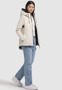 Veste imperméable beige clair avec capuche, accents noirs, fermeture éclair et poches latérales, portée avec un jean bleu clair et des baskets blanches.