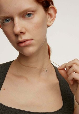 Collar de cadena de oro con un pequeño colgante que presenta un detalle brillante, llevado sobre un cuello desnudo, con un fondo neutro.