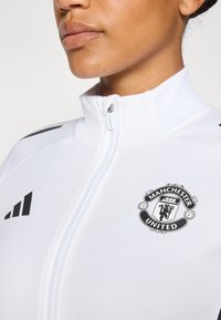 Veste blanche zippée arborant un logo de Manchester United en noir, avec des accents noirs sur les épaules. Fabriquée en matière lisse et légère.