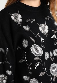 Pull noir en tricot avec une broderie florale blanche et grise complexe couvrant le devant et les manches.