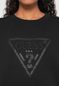Černá mikina s texturovaným trojúhelníkovým logem ve stříbrných kamínkách, s názvem značky "GUESS" a nápisy "WASHED" a "JEANS".