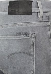 Vaqueros de mezclilla gris con una etiqueta de cuero negra, detalles de costura y un bolsillo trasero con una pequeña etiqueta de metal. Diseño clásico de cinco bolsillos.
