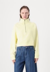 Gekürzter, Reißverschluss-Sweatshirt in Pastellgelb, mit hohem Kragen und Raglanärmeln. Kombiniert mit blauen, weiten Jeanshosen.