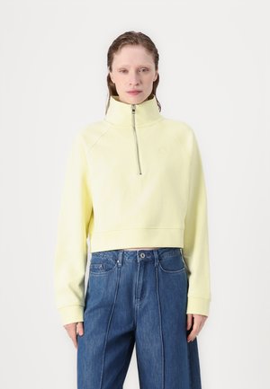 Cropped, zip-up sweatshirt in pastel yellow, featuring a high collar and raglan sleeves. Paired with blue denim wide-leg trousers.