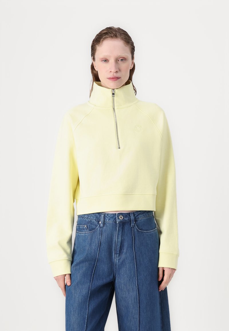 Gekürzter, Reißverschluss-Sweatshirt in Pastellgelb, mit hohem Kragen und Raglanärmeln. Kombiniert mit blauen, weiten Jeanshosen.