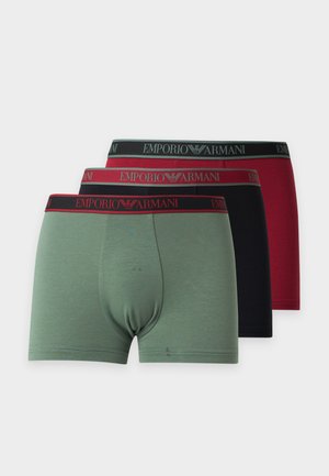 Skjorteløs mand iført sorte boxer shorts med rødt taljebånd står barfodet mod en ensfarvet hvid baggrund.