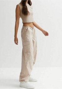 Béžový zkrácený top bez rukávů v kombinaci s oversized béžovými cargo kalhotami, které mají postranní kapsy a elastické manžety, doplněné bílými teniskami.