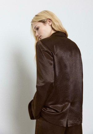 Blonde Frau im Seitenprofil, die eine glänzende dunkelbraune Anzugjacke vor einem schlichten hellen Hintergrund trägt.