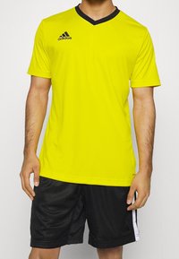 Camiseta deportiva amarilla de mangas cortas, con cuello en V y collar negro, que presenta el logo de Adidas en el lado izquierdo del pecho; combinada con pantalones cortos negros.