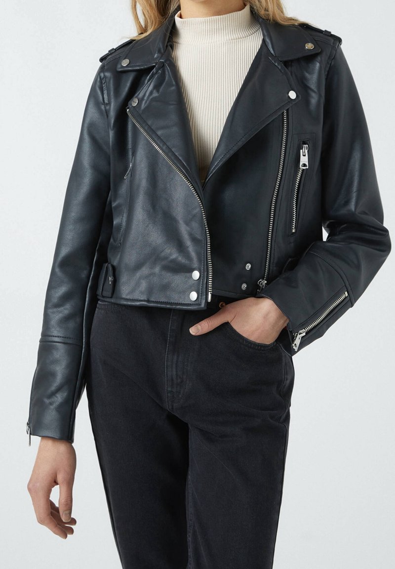 Personne portant une veste motard courte en cuir noir avec des fermetures éclair et des boutons argentés, associée à un pantalon noir et un col roulé côtelé beige clair.