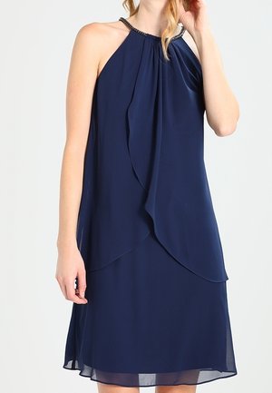 Woman wearing sleeveless navy blue halter dress with layered chiffon fabric and beaded neckline, posing with one hand touching her hair.