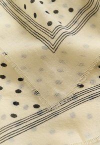 Tissu beige clair avec des pois noirs et des motifs de lignes parallèles, présentant une texture douce et pliée avec des bords effilochés.