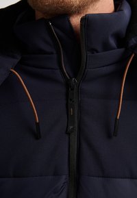 Chaqueta de marina con cuello alto, presenta una cremallera con detalles en negro y marrón, y paneles de tela texturizada.