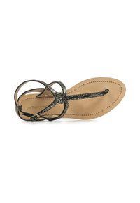 Sandali neri brillanti con suola piatta, caratterizzati da cinturini incrociati e un anello per le dita, posizionati su una soletta marrone chiaro con dettagli di cucitura.
