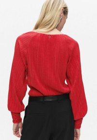 Rød plisseret bluse med rund halsudskæring, lange ærmer og knappede manchetter, kombineret med sorte bukser. Tekstureret stof med knappedetalje bagpå.