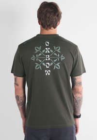 Camiseta de manga corta color verde oliva con un gráfico botánico en blanco y azul claro en la parte posterior, con cuello redondo y textura suave.