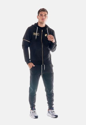 Jeune homme portant un sweat à capuche zippé noir avec un patch "1947", un pantalon de jogging noir et des baskets blanches, debout devant un fond clair uni.