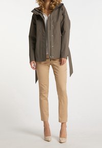 Veste imperméable kaki avec capuche, fermeture éclair à l'avant et pressions, associée à un pantalon beige slim et des talons de couleur claire.