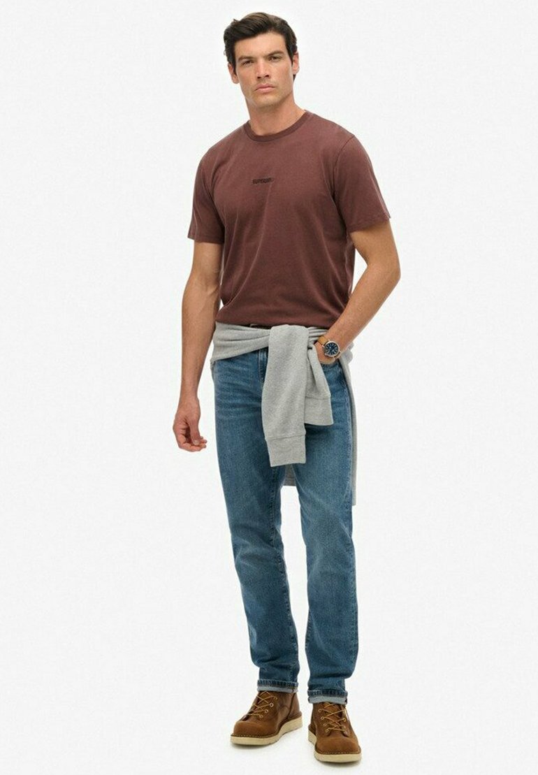 Brązowa koszulka z okrągłym dekoltem, jasnoszary sweter zarzucony na talię, niebieskie jeansy, brązowe buty z żółtymi akcentami, casualowy strój.