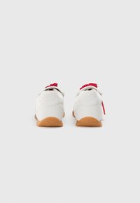 White sneakers with a textured upper and red laces. Rubber sole with a herringbone pattern. Branding visible on the heel.