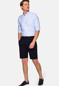 Jasnoniebieska koszula zapinana na guziki z podwiniętymi rękawami, zestawiona z czarnymi szortami. Model nosi ciemne sneakersy. Czysty, minimalistyczny design, casualowy wygląd.