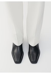 Černé kožené kotníkové boty se čtveratým špičkou a hladkým povrchem, spárované s bílými zúženými kalhotami, pohled shora.