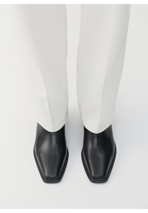 Botas de cuero negras hasta el tobillo con punta cuadrada y textura lisa, combinadas con pantalones blancos ajustados, vistas desde arriba.