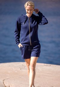 Navy blue zip-up hoodie with a front logo and matching shorts. Material appears soft and comfortable. Features side pockets and drawstring waist.