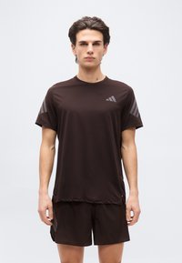 Νέος άνδρας που στέκεται φορώντας σκούρο καφέ αθλητικό μπλουζάκι και ταιριαστό σορτς με διακριτικές ρίγες λογότυπου, ουδέτερο φόντο.