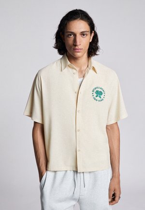 Jonge man met schouderlang haar draagt een crème kortärmelig overhemd met knopen en groen "Sports Legacy New York"-logo en lichtgrijze broek.