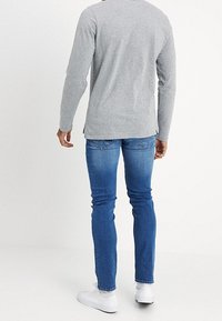 Man som står med ryggen vänd mot kameran, iklädd en grå långärmad skjorta, blå slim-fit jeans och vita sneakers, mot en enfärgad vit bakgrund.