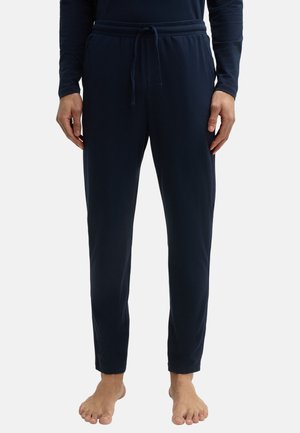 Mężczyzna ubrany w granatowe spodnie z sznurkiem i bluzkę z długim rękawem, stojący boso na jednolitym tle.